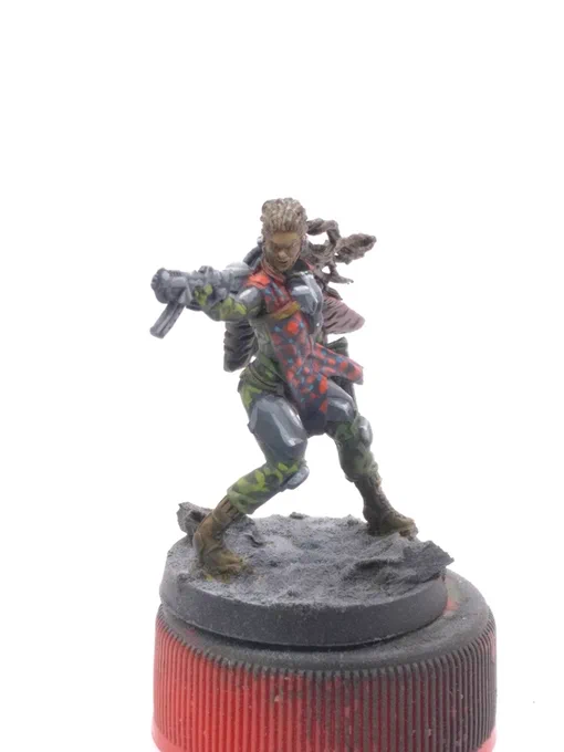 Aida Swanson from Corvus Belli's Infinity Defiance boardgame. humanoid woman with dark skin and braids brandishing a pair of submachineguns. green fatigues, gunmetal armor plates, and a red and blue plaid pattern resembling Maasai shuka cloth.