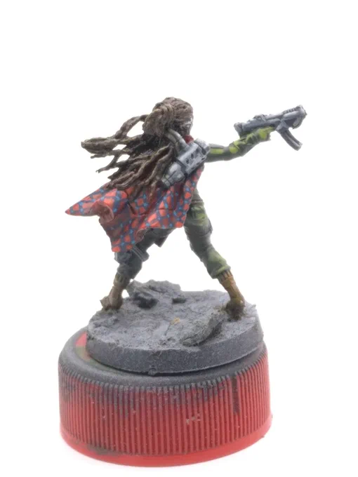 Aida Swanson from Corvus Belli's Infinity Defiance boardgame. humanoid woman with dark skin and braids brandishing a pair of submachineguns. green fatigues, gunmetal armor plates, and a red and blue plaid pattern resembling Maasai shuka cloth.