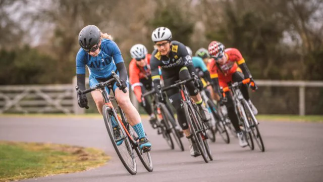 The image shows a group of women racing road bikes on the tarmac surface at the odd Down Sports Ground. 