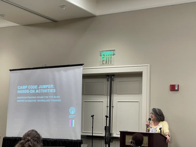 Camp Code Jumper: Hands on Activities title on presentation board, woman speaking at podium