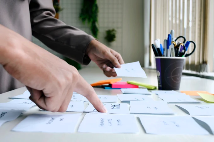An image of the hands of two people pointing to post-it notes on a desk.