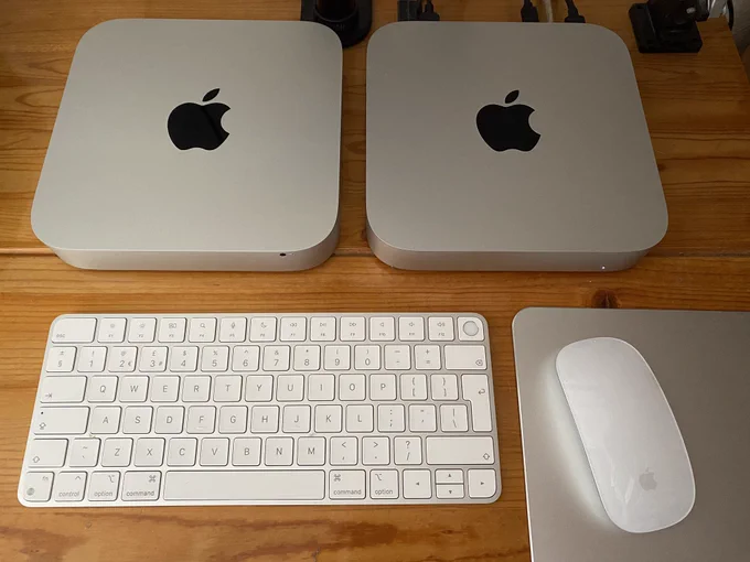 A pair of Mac Minis from above, sitting on a desk, with a keyboard and mouse in front of them. The one on the right appears to have leads connected at the back of it and its power light is on. The one on the left looks otherwise identical.