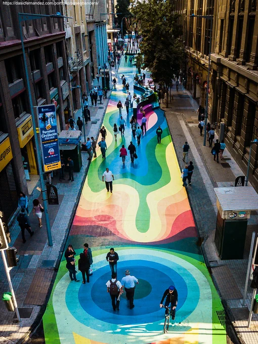 image of Paseo Bandera in Santiago Chile. Street is closed to cars and open to people. Painted with a mural that is said to relate to indigenous heritage, which looks like bold rainbow color gradients in different shape. Places to sit. Public restroom. Retail along the side. trees. people enjoying the space