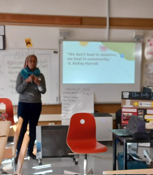 An photo of Paula standing at the front of a classroom with a slide that says "we don't heal in isolation, we heal in community." 