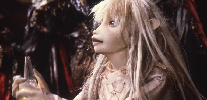 Kira the Gelfling from The Dark Crystal. She is pale and gaunt; her clothes, skin, and hair are all approximately the same shade of almost-white beige. She holds the Crystal Shard in her hands and is surrounded by Skeksis.