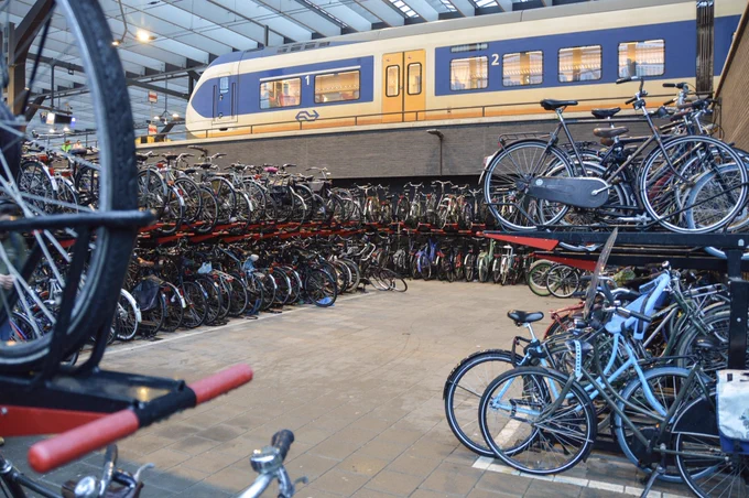 A bicycle parking facility at Rotterdam Centraal Station is filled with dozens of bikes on two-tiered racks. A blue and yellow train waits at the platform above.
