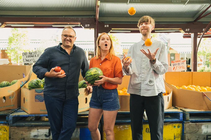 Photo of new Triple R Breakfasters team. Daniel, Nat and Simon are standing in a row at a fruit stall at the markets. Daniel is smiling at the camera while Nat's got a shocked look... as Simon juggles some oranges! Image credit: Naomi Lee Beveridge.