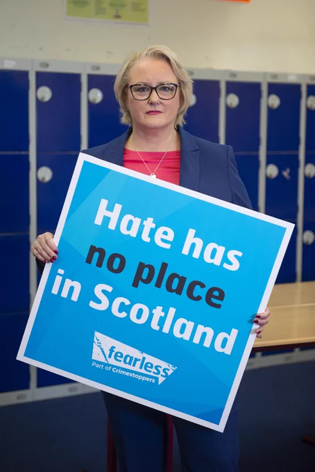 Minister for Equalities Christina McKelvie stands in front of school lockers wearing navy suit holding blue square sign that reads 'hate has no place in Scotland 