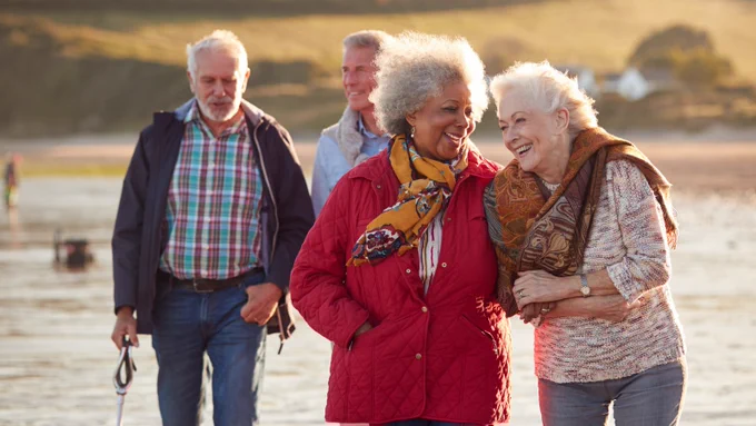 Photo of 2 pairs of elderly men and women wearing jackets and scarves taking a walk along a water's edge. The two women in the foreground are linking arms.