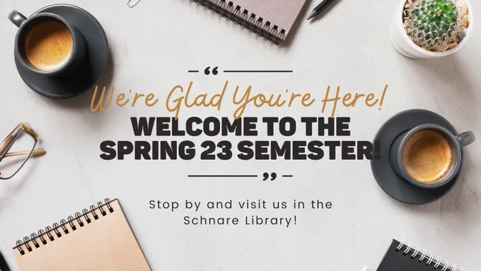 A warm gray table is covered with two dark grey mugs filled with coffee, three top spiral notebooks, a pair of glasses, a pencil, and a small cactus. The text reads: We're glad you're here! Welcome to the Spring 23 Semester! Stop by and visit us in the Schnare Library!