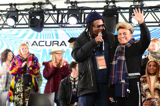 Rodger Ross Williams, a Black man with long dark dreads, and Cassandro, a Mexican man with short blond hair, stand and wave to an audience at Outfest Outpost.