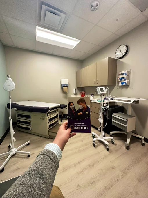 David O.’s left arm extends from the bottom left corner holding a brochure titled “Welcome to the Glenbrook School Health Center” as he sits in a chair in the corner of an exam room. In the background are a light’ exam table, counters, vital checker cart, and care provider’s mobile standing desk.