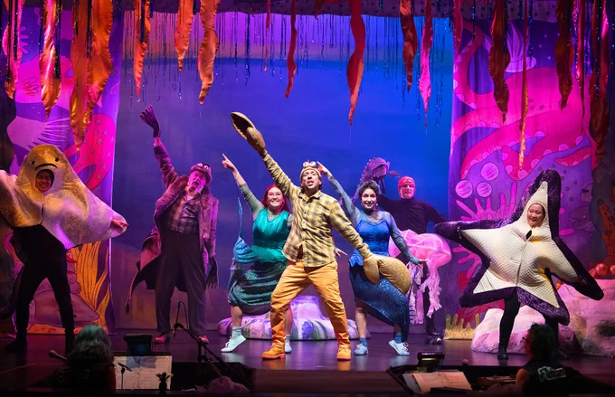 Production photo from East Van Panto: The Little Mermaid of the cast dancing onstage. All seven actors onstage are wearing colourful costumes and the set is decorated as a vibrant underwater scene.