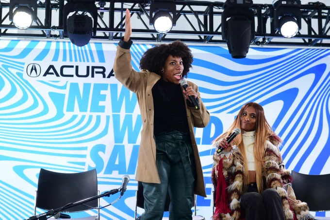 Y-vonne Hutchinson, a Black woman with short dark hair, and D Smith, a Black woman with long brown hair with blond highlights, greet the audience at Outfest Outpost.