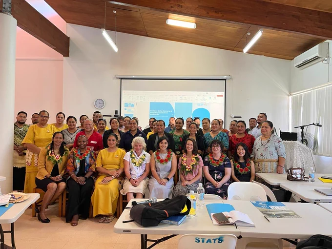 A large group of people gathered for a photo. Many of the people are Tongan and some are wearing lei around their necks. Many people are wearing yellow. They are indoors and standing in front of a screen with a presentation. 