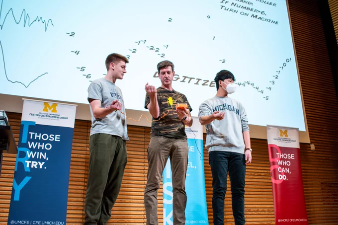 Two students stand next to Greg Gage. They have orange cables attached to their arms. 
