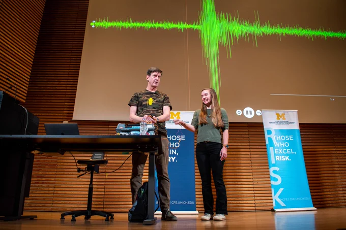 A young woman with long hair stands next to Greg Gage. She has orange cables attached to her arm that are connected to a box. In the background, a visual indication of her heart and brain activity is displayed as a green graph.