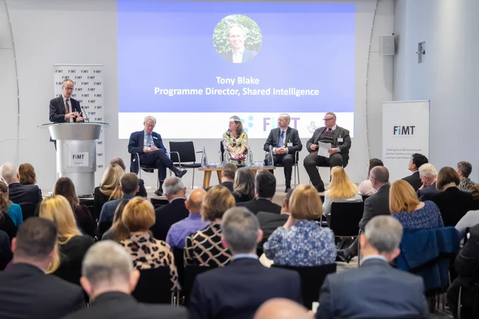 A panel of people, with one man standing and speaking, in front of an audience. A powerpoint slide reads: Tony Blake, Programme Director, Shared Intelligence 