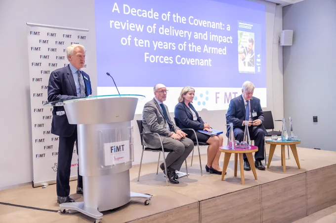A panel of people on a stage, with one man standing up and speaking into a microphone. The slide behind them reads: A Decade of the Covenant: a review of deliver and impact of ten years of the Armed Forces Covenant