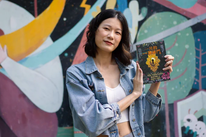 East Asian girl with short, curly hair, in a jean jacket, standing in front of a painted wall and holding a traditional Chinese copy of "the ten thousand doors of January"