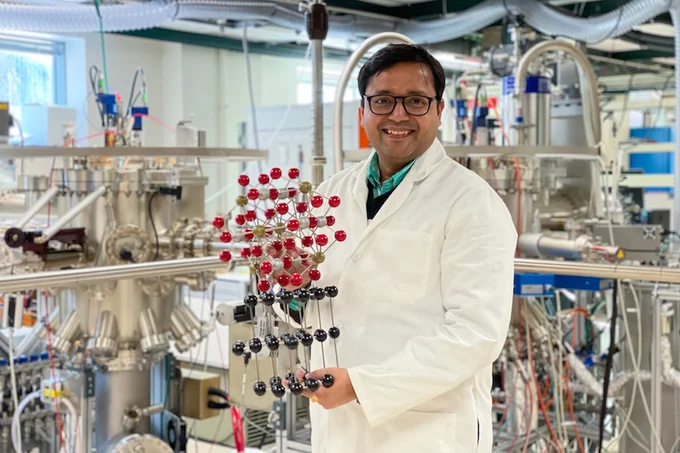Photo of Bharat Jalan (a Southeast Asian man in a lab coat) holding a molecular model, standing in a lab full of equipment