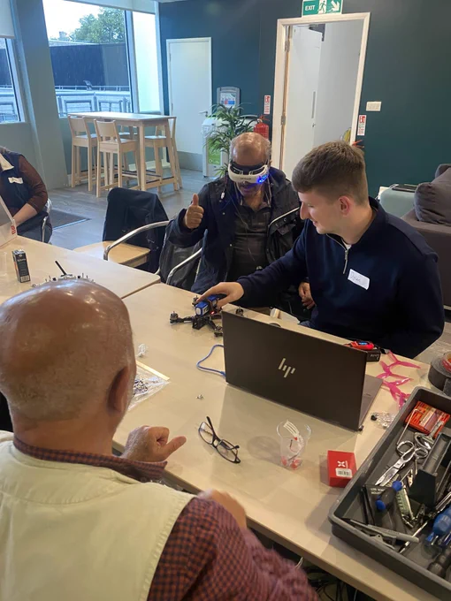 A group of men sitting round a table with a laptop, one of the men is wearing a VR headset 
