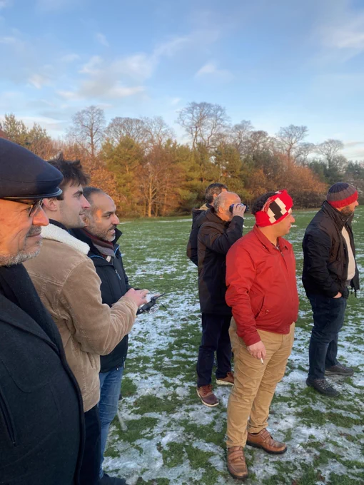 A group of men standing outside in a park with one of the men holding a drone remote 