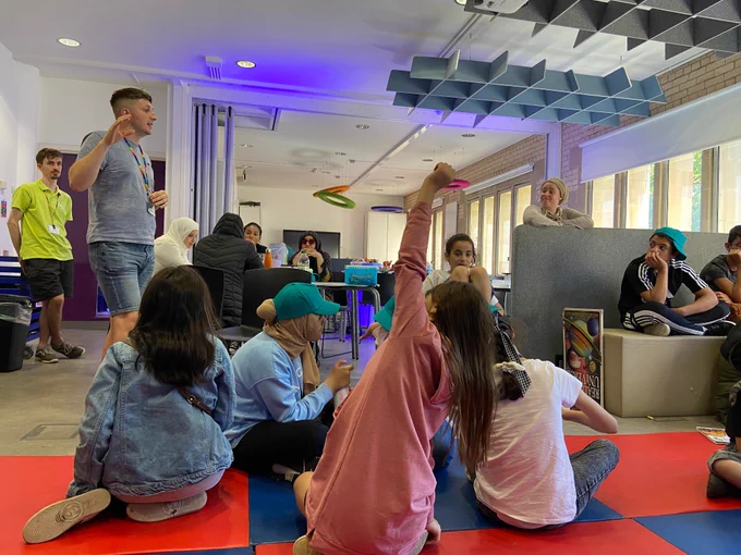 Group of children sitting on the floor as a man gives a talk, one of the children has their hand up to ask a question