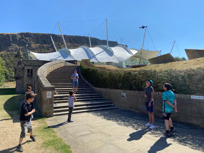Man standing on stairs flying a drone with a group of kids at the bottom of the stairs watching the drone 