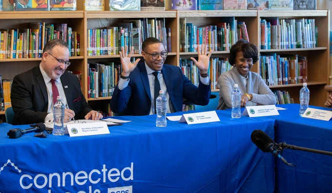 Secretary Miguel Cardona meeting with several others inside a school library. Man in center has arms up and smiling/laughing