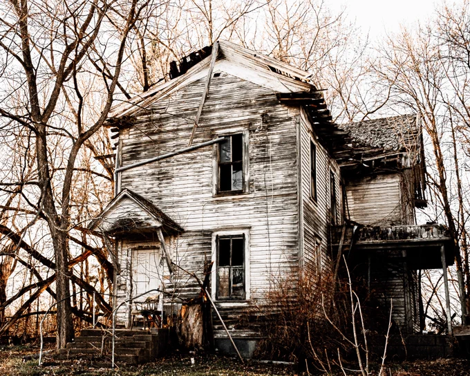 An overexposed photo of a decaying old house.  There are bare trees around it. The sky is white.