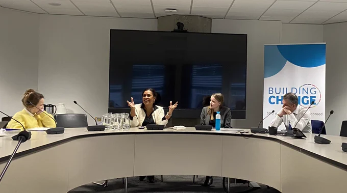Vier mensen in gesprek aan een ovalen tafel. Banner met “Building Change” op de achtergrond.