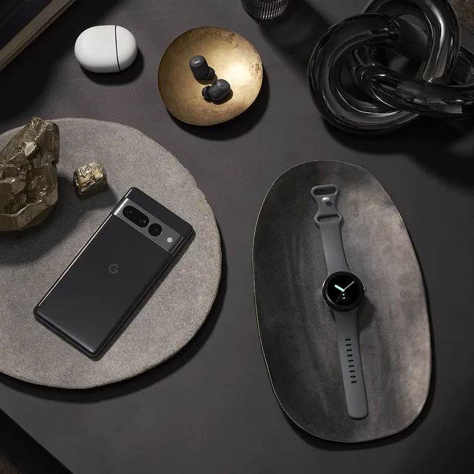 The Google Pixel 7 Pro in Obsidian, Pixel Buds Pro in Charcoal, and Pixel Watch in Polished Silver case / Charcoal Active band displayed on various dishes across a black surface.