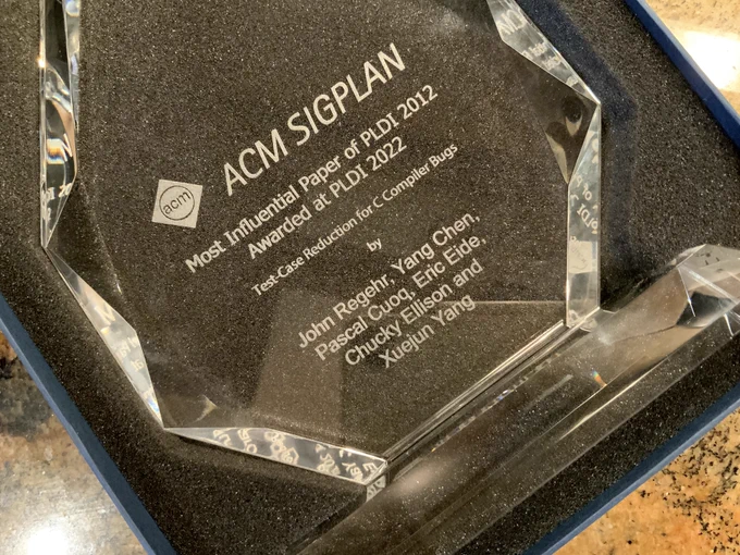 Photograph of a glass award in box. Text: "ACM SIGPLAN / Most Influential Paper of PLDI 2012 / Awarded at PLDI 2022 / Test-Case Reduction for C Compiler Bugs / by / John Regehr, Yang Chen, Pascal Cuoq, Eric Eide, Chucky Ellison and Xuejun Yang"