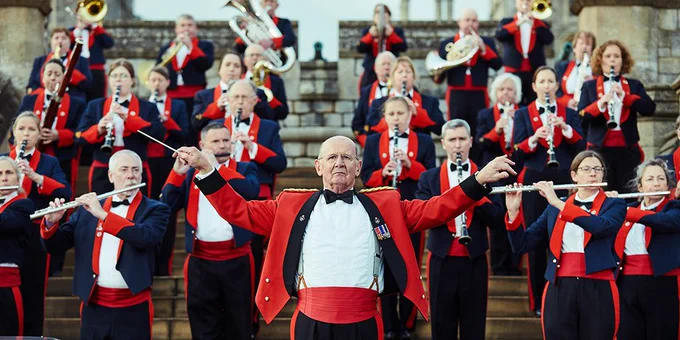 The Central Band of the Royal British Legion are pictured on a set of steps at Windsor Castle. They are wearing their uniforms of navy jackets with red lapels with black trouser with red stripes down the sides. David Cole stands in the centre wearing a red jacket with black lapels and raising his hands as if to conduct wiht his baton. The band are picture with a variety of their instruments, including drums, flutes, clarinets, trumpets and cymbal.