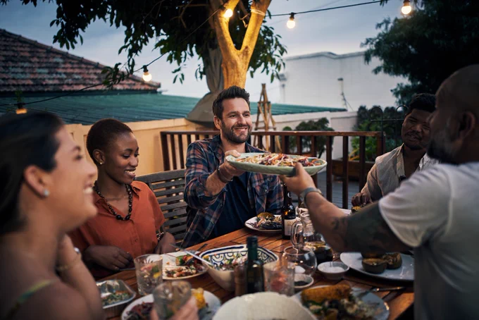 A table with people gathered around, sharing food. Two men are passing a plate between them.
