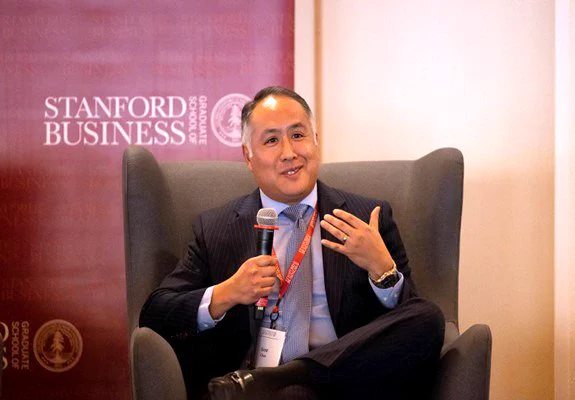 Scott Chan speaks, sitting in a chair. He has short brown and gray hair and is wearing a suit jacket, blue shirt and tie. He's holding a microphone.