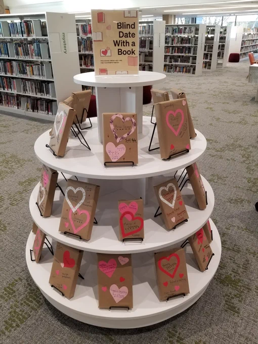 White display shelving with a sign on top saying "Blind Date With a Book." The shelves are filled with books wrapped in brown paper, decorated with red, pink, and white hearts and descriptive words.