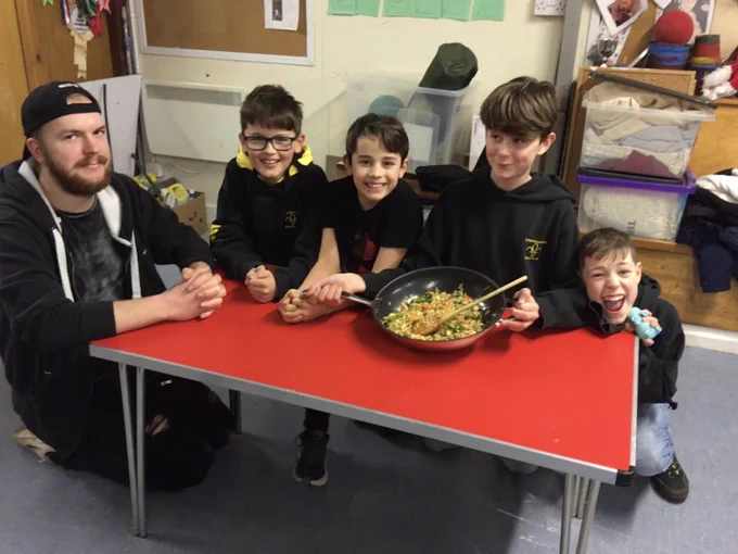 4 Scouts & a leader wearing black tops kneeling around a red table that has a wok of colourful rice on it.