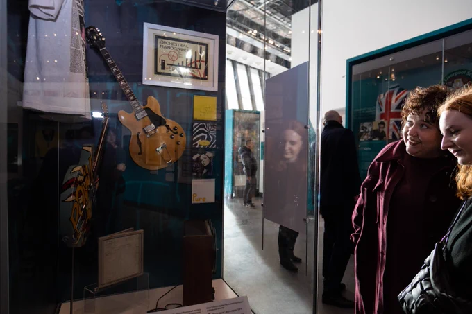 Two visitors in Wondrous Place gallery at Museum of Liverpool. 
Two visitors, to the right of the image, are looking at a glass case with objects inside. Objects include two guitars, framed pictures and a t-shirt. In the background other visitors are visible with their backs to the camera, looking around the rest of the gallery. Image credit: Robin Clewley.