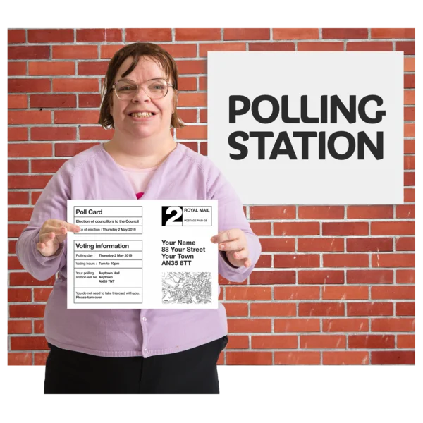 A woman holding a poll card in front of her. She has dark hair, glasses and a pink top. Behind her is a brick wall with a white poster with black text that says POLLING STATION.