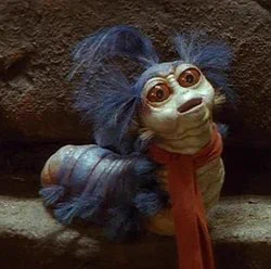 William the Worm from Labyrinth. He is a small blue and white worm with three large tufts of blue hair on his head and small tufts of blue hair along his side. He has large red eyes and wears a red scarf.