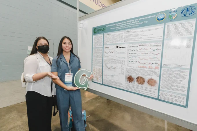 Two women stand smiling in front of a research poster. One woman holds a large letter "G" cutout.