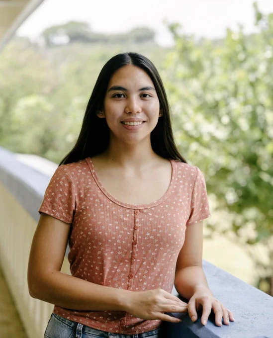 A woman with long black hair wearing a pink button-down short-sleeved shirt with a pattern of small white feathers. She is smiling and standing next to a railing with trees and foliage in the background.