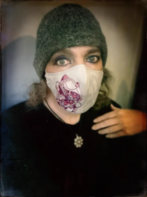 Selfie. SheFaerer, a white woman, the fingers of her left hand touching her left shoulder area. She wears a black top, a silver pendant with rhinestones on a black cord, a silver-gray toque, and a gray mask with a mecha kitty in purples, black, and white. She looks into the camera, her eyes done in heavy black eyeliner and mascara, with a blend of purples and rose and shimmer eyeshadows.