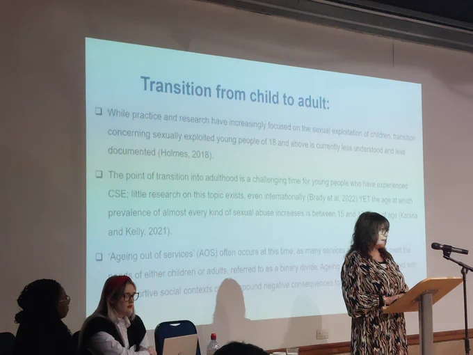 A woman (Professor Brady) presents research findings from a slide on a large screen. Two woman (Geraldine Brown and Bailey Foster) sit at a table alongside  and contribute to information sharing.