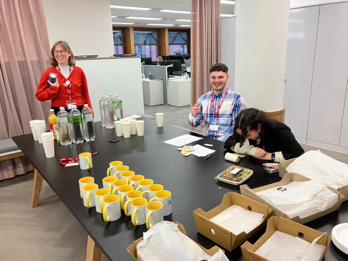 Three members of the We and AI team prepare drinks, name badges and food 