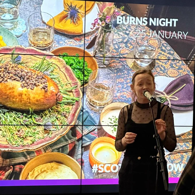 A photograph of Clare Campbell stood in front of a digital backdrop of a Burns dinner table