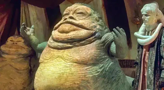 Jabba the Hutt from Star Wars. He is a large, slug lake alien with wrinkly green skin, a huge mouth, and wrinkly eyes. He is flanked by another Hutt and a humanoid alien with white skin and a long tentacle coming from their head.