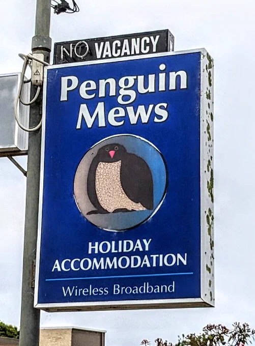 A blue hotel sign for Penguin Mews. The sign features a stylised image of a pengion in the middle. The image is a bit fatter than typical and looks a bit like an owl.